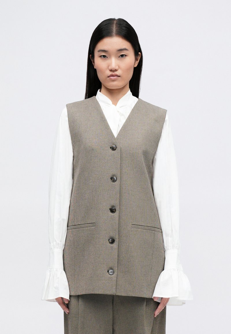 Γκρίζα αμάνικη γιλέκα με υφαντή υφή, πέντε κουμπιά μπροσ�τά και δύο πλαϊνές τσέπες, σε συνδυασμό με μια λευκή μπλούζα.