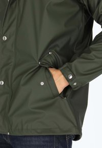 Groene waterdichte jas met een gladde textuur, drukknopen, zijzakken en verstelbare manchetten. Het ontwerp benadrukt functionaliteit en duurzaamheid.