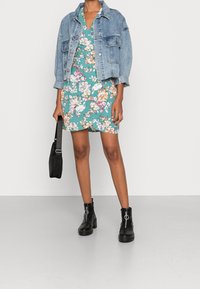 Vestido floral en verde azulado, chaqueta vaquera, bolso negro y botines. Con mangas cortas, detalle de cremallera y ajuste casual.