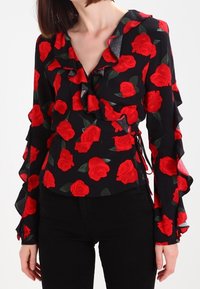 Blusa nera con maniche a volant e stampa di rose rosse, dotata di design avvolgente e laccetti laterali regolabili. Texture del tessuto liscia.