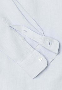 Poignets de chemise blancs avec détails de bouton et de boutonnière, montrant une partie de la manche repliée et les coutures sur un fond de tissu clair.