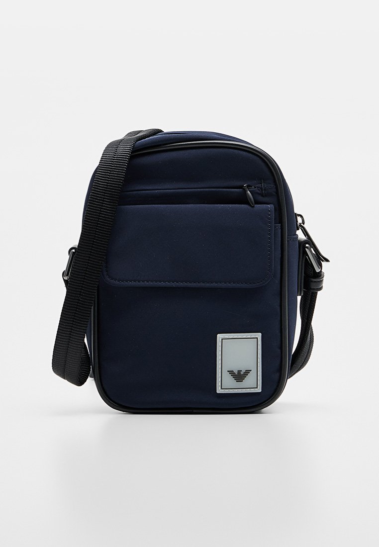 Bolso cruzado color azul marino fabricado con tela duradera. Cuenta con un bolsillo frontal, correa ajustable y una etiqueta con logo en la parte delantera. Cierre con cremallera.