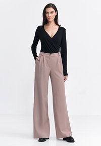 Czarna bluzka z długim rękawem w stylu kopertowym, zestawiona z szerokimi, beżowymi spodniami, posiadającymi zapięcie na guziki i kieszenie, na neutralnym tle.