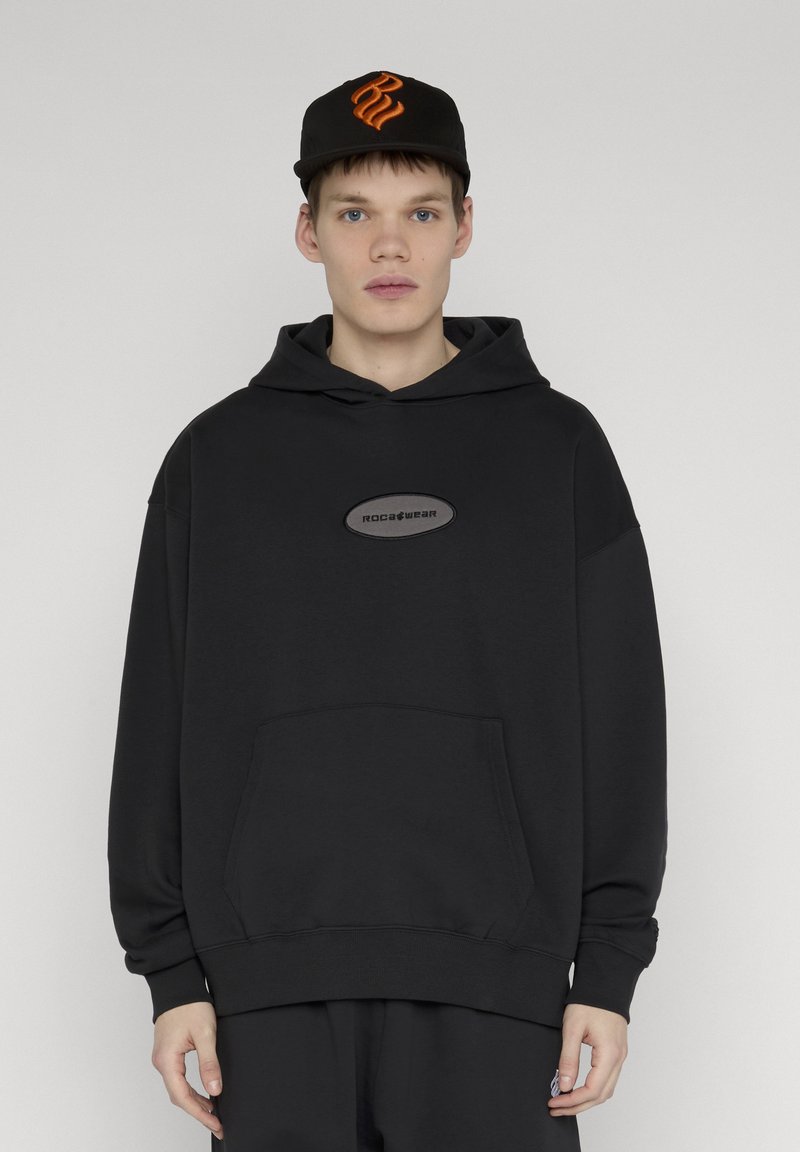 Jeune homme portant un sweat à capuche noir avec le logo "Rocawear" et une casquette noire avec un emblème orange, debout devant un fond clair uni.