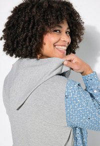 Sweatshirt à capuche grise vu de dos avec des manches embellies de sequins bleus. Caractéristiques d'un tissu texturé, d'une coupe décontractée et de coutures visibles.