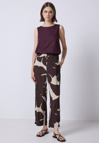 Mouwloze bordeauxrode top, gecombineerd met brede, bruine broek met een crèmekleurig bladerenpatroon. Het model toont een ontspannen pasvorm en zijzakken.