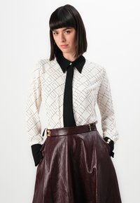 Žena se rovnými tmavými vlasy má na sobě bílou vzorovanou blůzu s černým límečkem a manžetami, kterou doplňuje burgundskou koženou sukňou s vysokým pasem.