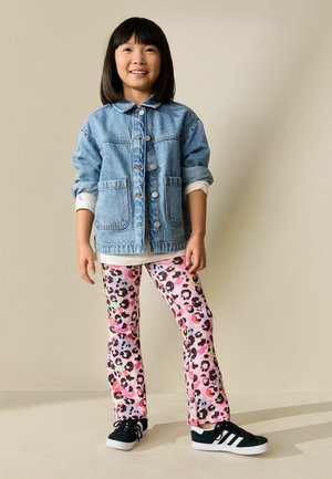 Young girl smiling, wearing a blue denim jacket, colorful leopard-print pants, and black sneakers with white stripes, standing against a beige wall.