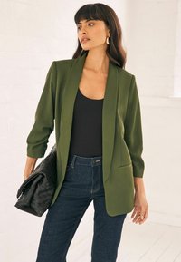 Blazer verde oliva con un diseño entallado, solapas con muesca, mangas largas y almohadillas en los hombros, combinado con una blusa negra y jeans oscuros.