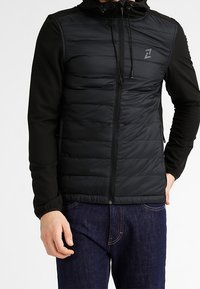 Man i svart vadderad jacka med huva, dragkedja fram och mörkblå jeans, står med en hand i fickan.