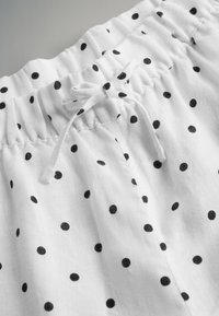 Bela tkanina z majhnimi črnimi pikami. Ima elastičen pas z zavezovalnim vrvico. Mehka tekstura in lahka zasnova.