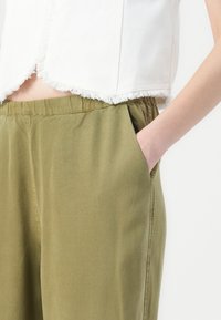 Kuttet hvit denimtopp med frynset kant og olivengrønn bukse med elastisk midje og sidetasker. Glatt, avslappet stofftekstur.