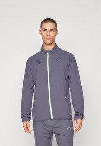 Graue Sportjacke mit hohem Kragen, fluoreszierendem grünen Reißverschluss, schwarzen Logo-Prints und Seitentaschen. Das Material wirkt leicht und strukturiert.