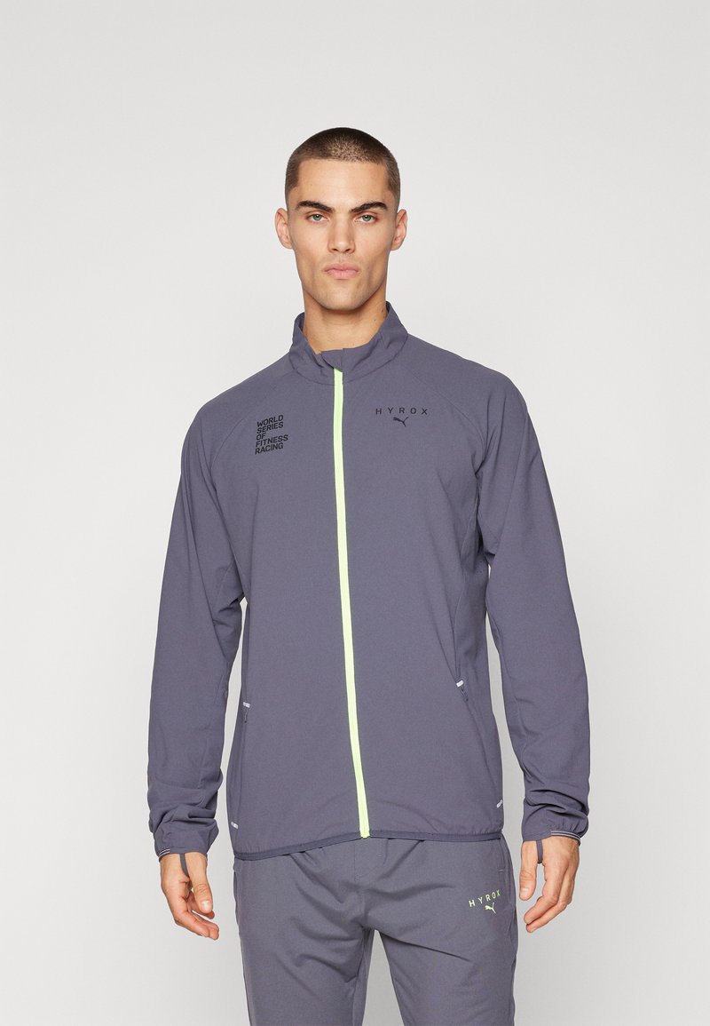Graue Sportjacke mit hohem Kragen, fluoreszierendem grünen Reißverschluss, schwarzen Logo-Prints und Seitentaschen. Das Material wirkt leicht und strukturiert.