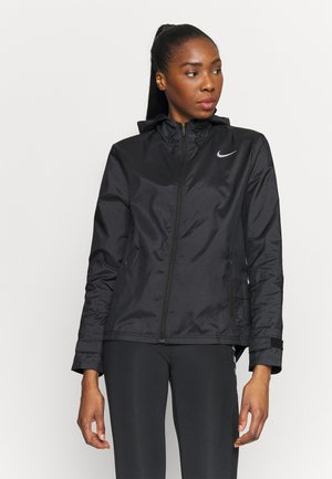 Kobieta ubrana w czarną kurtkę na zamek Nike i czarne legginsy, stojąca na tle jednolitego jasnego tła, patrząca w bok.