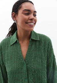 Camicia button-up verde, strutturata e con colletto, caratterizzata da un aspetto stropicciato e grandi bottoni turchesi.