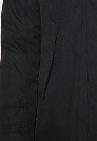 Černá zateplená bunda s měkkou texturou, která má boční kapsu, strukturovaný design a viditelné stehům.