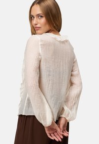 Lightweight, sheer cream blouse with a textured finish, ruffled neckline, and elasticized sleeves. Features a relaxed, loose fit.