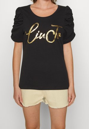 Zwart T-shirt met korte mouwen en opgeblazen mouwen, met metallische gouden tekst "linJo"; gedragen met lichtgekleurde versleten denim shorts.