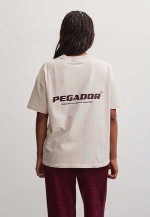 Persoon met vlechtjes draagt een beige t-shirt met de tekst "PEGADOR" op de rug en bordeauxrode broek, staand tegen een effen achtergrond.
