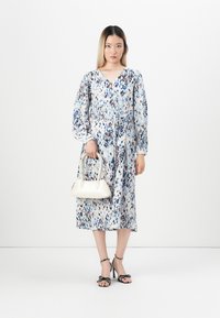 Woman in a long-sleeve, knee-length speckled dress holding a white handbag, wearing black high-heeled sandals with bow accents.