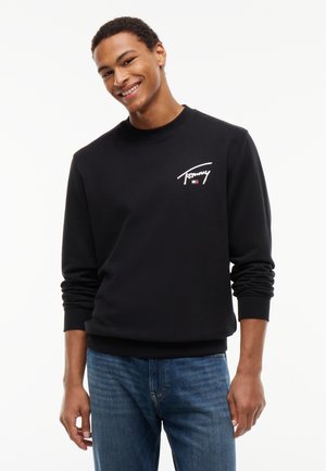 Jeune homme souriant portant un sweat-shirt noir Tommy avec logo sur la poitrine et un jean bleu, debout devant un fond blanc uni.