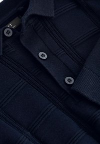 Marinblå stickad polo med ett texturerat mönster, försedd med tre svarta knappar och en krage. Slät och mjuk tyg.