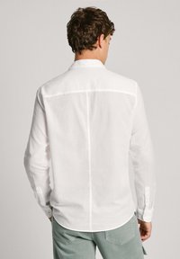 Camisa blanca de manga larga hecha de lino, con cuello abotonado, dobladillo recto y costuras sutiles en la parte posterior. Combinada con pantalones verde claro.