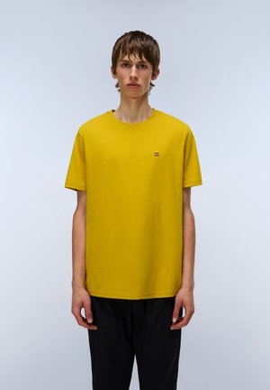 Yellow cotton t-shirt with a round neckline and short sleeves, featuring a small embroidered emblem on the chest. Worn with black pants.