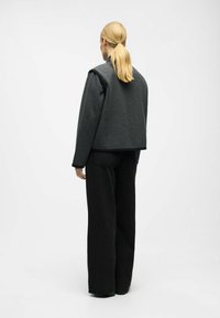 Woman with blonde ponytail wearing a dark gray jacket and black wide-leg pants, standing and facing away against a plain background.