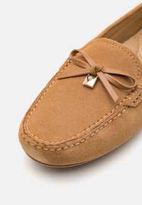 Mocassin en daim beige avec détails cousus, un nœud en cuir noué et un petit charm carré doré sur la partie supérieure avant.