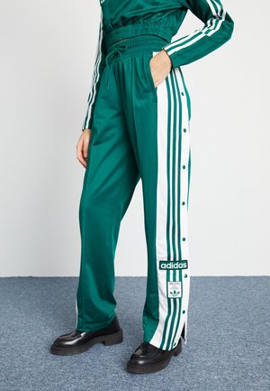 Osoba nosząca zielone dresowe spodnie Adidas z białymi paskami po bokach i zatrzaskami, w połączeniu z czarnymi mokasynami na niebieskim dywanie.