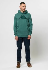 Zelená mikina s kapucňou a čiernym logom na hrudi, s kangaroo vreckom a rebrovanými manžetami, spárovaná s tmavými džínsami a hnedými čižmami.