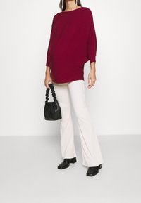 Femme portant un pull ample rouge foncé, un pantalon blanc évasé, des bottines noires à texture, et tenant un sac à main noir à poignées froncées.