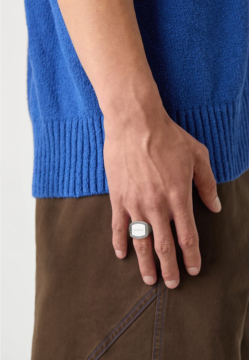 Silver ring with rectangular face, featuring engraved brand name. Hand resting against blue textured sweater and brown pants.