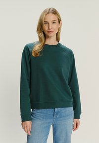 Žena s blond vlasy, která má na sobě zlaté kruhové náušnice, tmavě zelenou mikinu s dlouhým rukávem a světle modré džíny, proti jednobarevnému pozadí.