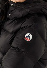 Giacca a vento nera con finitura lucida, dotata di colletto alto, design trapuntato e un patch circolare con logo sulla manica sinistra.