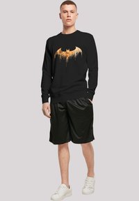 Fekete pulóver narancssárga csepegő Batman logóval, kerek nyakkal, hosszú ujjú, fekete rövidnadrággal és fehér sportcipővel párosítva.