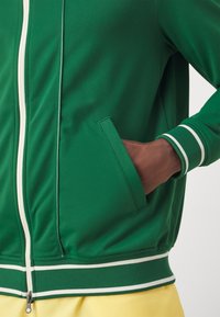 Groene sportjack met witte rits, zijzakken en gestreepte manchetten. Gladde textuur met geribbelde zoomdetails. Gele shorts op de achtergrond.