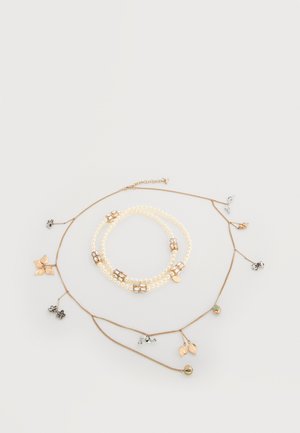 Trois colliers superposés avec des perles de culture, des accents en métal doré et des breloques en forme de feuille avec différentes finitions, disposés sur un fond clair.