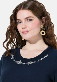 Námořnické tričko s bílou vyšitou rybkou a listovými motivy podél výstřihu. Modelka má na sobě zlaté a tkané náušnice a předvádí texturované vlasy.