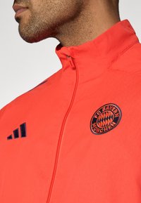 Κόκκινο τζάκετ με φερμουάρ, κατασκευασμένο από ελαφρύ ύφασμα. Διαθέτει ψηλό γιακά, ναυτικό μπλε λεπτομέρειες και κεντημένο λογότυπο FC Bayern München στο στήθος.