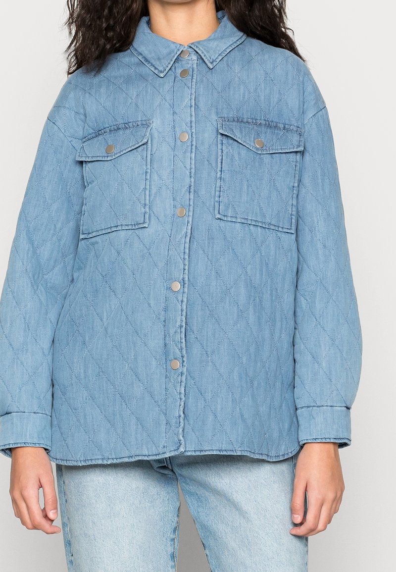 Femme portant une veste en jean matelassée bleu clair avec des boutons-pression et deux poches à rabat à l'avant, portée sur un jean bleu clair.