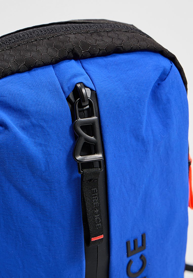 Blauer Rucksack mit oberer Fläche im schwarz-hexagonalen Muster, schwarzen Beschlägen und einem auffälligen Reißverschluss. Verfügt über einen Trageriemen mit dem Text "FREERIDE".