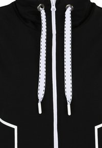 Schwarzer Kapuzenpullover mit einem Reißverschluss vorne, weißen Akzenten und dicken weißen Kordeln mit schwarzen Perforationen. Weicher Stoff mit glatter Textur.