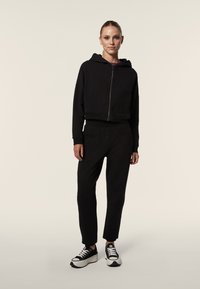 Sudadera negra corta con cierre de cremallera, combinada con pantalones negros ajustados y zapatillas negras. Textura suave, diseño minimalista.
