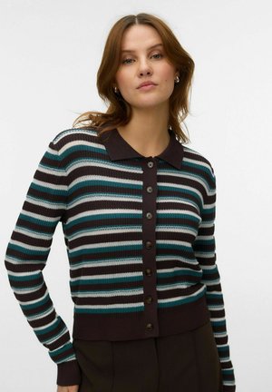 Femme portant un cardigan rayé boutonné avec des rayures horizontales noires, bleu sarcelle et blanches ainsi qu'un col foncé sur un fond uni.