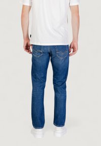 Jeans in denim blu con una vestibilità dritta, caratterizzati da una cucitura gialla sulle tasche posteriori, indossati con una maglietta bianca e sneaker bianche.