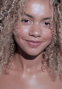 Ung person med lys brun hud og krøllete blondt hår som smiler forsiktig, med synlige skuldre og glødende hud.