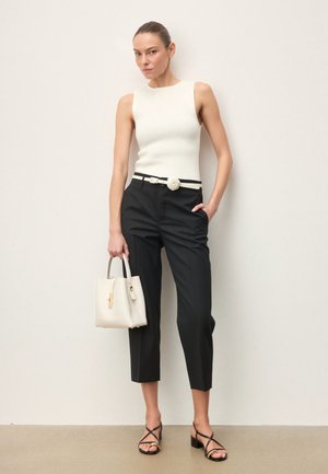 Frau trägt weißes ärmelloses Oberteil, schwarze verkürzte Hose mit weißem Gürtel, hält weißen Handtasche und trägt schwarze Riemchensandalen, steht an einer Wand.